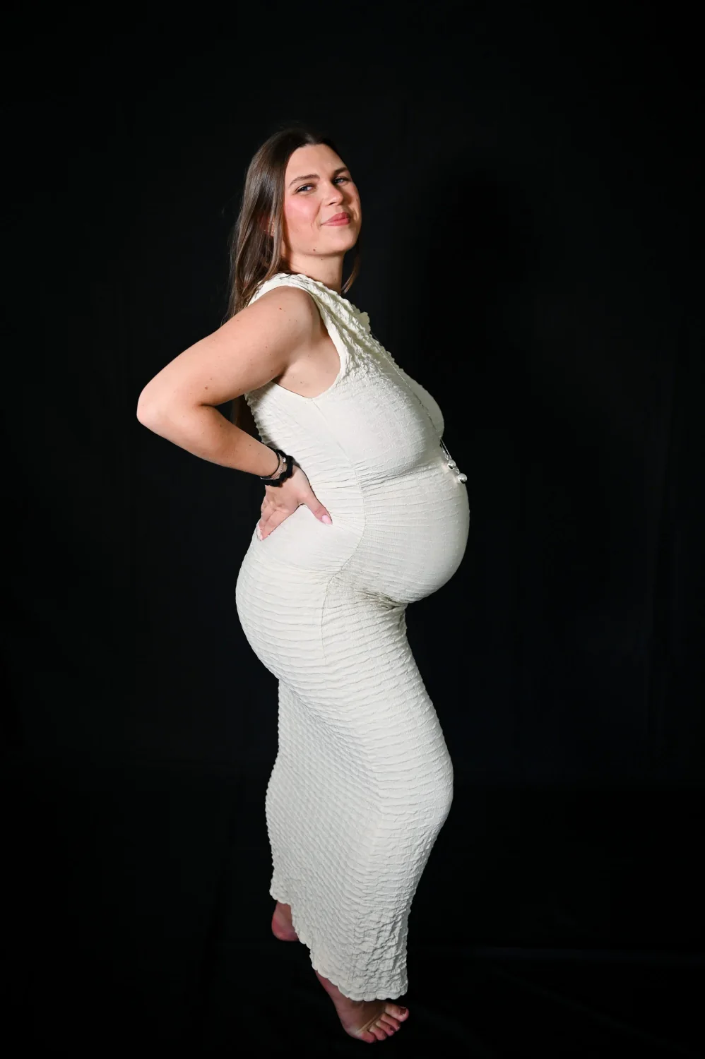Future maman en robe blanche moulante sur fond noir, photo de grossesse en studio